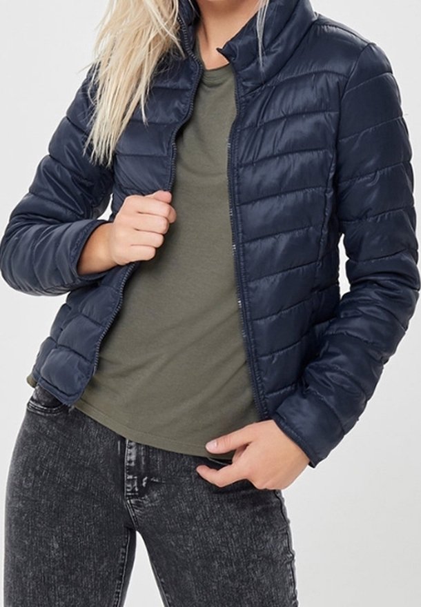 Giacca puffer navy con design trapuntato, colletto alto e zip frontale. Indossata sopra una t-shirt oliva, abbinata a jeans scuri. Texture liscia.