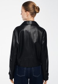 Giubbotto in pelle nera con colletto, design corto e polsini con bottone. Texture liscia con lucentezza sottile, abbinato a jeans blu.