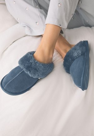Pieds portant des chaussons bleus pelucheux et un pantalon de pyjama gris clair avec de petits motifs de pingouins reposant sur un lit blanc.