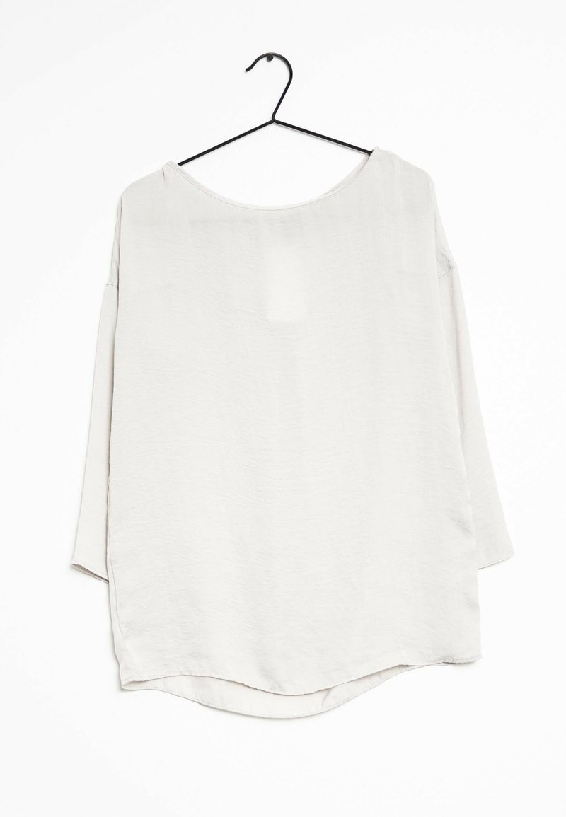 Blouse légère grise, ample, à manches longues en tissu texturé. Dispose d'un encolure large et d'un ourlet arrondi. Suspendue à un crochet noir.
