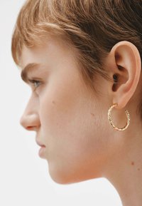 Pendiente de aro dorado retorcido mostrado en la oreja de una persona con cabello corto, con una textura lisa y un acabado brillante.