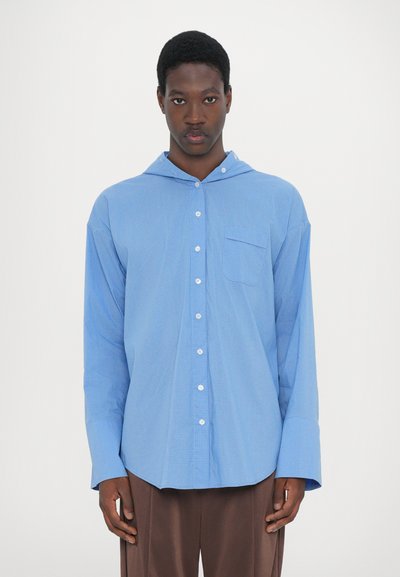 Chemise boutonnée bleu clair avec capuche, dotée d'une petite poche sur la poitrine et d'un motif à carreaux. Manches longues avec larges poignets.