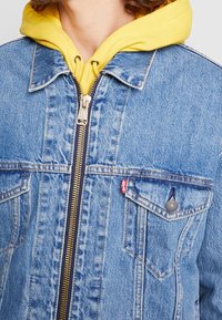 Jeansjacka med en guldfärgad dragkedja, två bröstfickor med logodetaljer och en gul hoodie som tittar fram under.