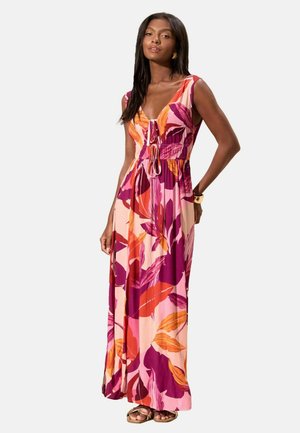 Kvinde iført en ærmeløs maxi kjole med V-hals og store blomster i pink, lilla, orange og rød, stående med den ene hånd på hoften.