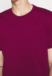 Primer plano de una persona que lleva una camiseta de cuello redondo, de manga corta y color púrpura oscuro, que muestra el cuello, los hombros y la parte superior del pecho.