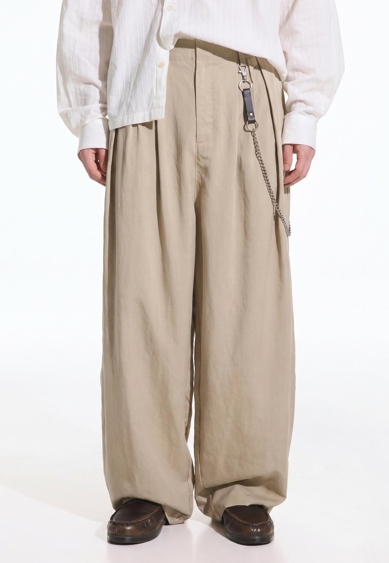 Personne portant un pantalon large beige avec un porte-clés en cuir noir et une chaîne argentée, associé à des chaussures marron foncé et une chemise blanche à manches longues.