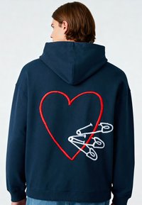 Felpa blu navy con un grande cuore rosso ricamato e spille da balia bianche sul retro, dotata di tasca a marsupio e cappuccio con coulisse.