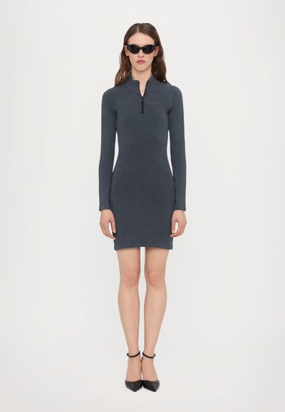Rochie lungă de culoare gri închis, cu guler cu fermoar, formă ajustată și textură netedă, asociată cu tocuri stiletto negre și ochelari de soare.