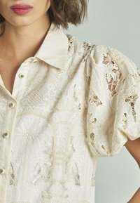 Cremfarbene Bluse mit aufwendiger Bestickung, einem kragenförmigen Ausschnitt und puffartigen Ärmeln. Der Stoff weist spitzenartige Ausschnittmuster auf.