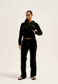 Melna velūra sporta tērps ar aizdari kapuci un saistāmām biksēm. Iespējas ar logotipa detalizāciju un īsu dizainu. Izskatu papildina čības.