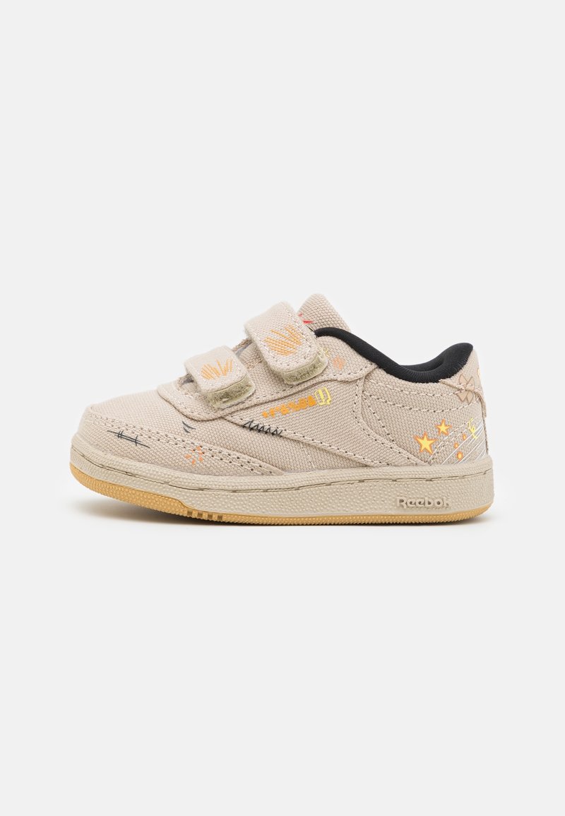 Zapatilla de lona beige con dos tiras de Velcro, forro negro y detalles bordados multicolor que incluyen estrellas y líneas a lo largo de los lados.