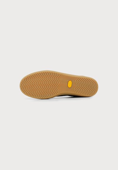 Semelle de chaussure avec motif en caoutchouc beige en zigzag et un petit logo jaune près du centre, sur un fond gris clair.