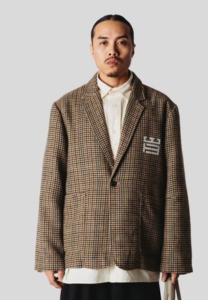 Hombre con el pelo largo recogido en una coleta baja, vestido con un blazer marrón de cuadros oversized sobre una camisa color crema, sosteniendo una bolsa beige sobre un fondo liso gris claro.