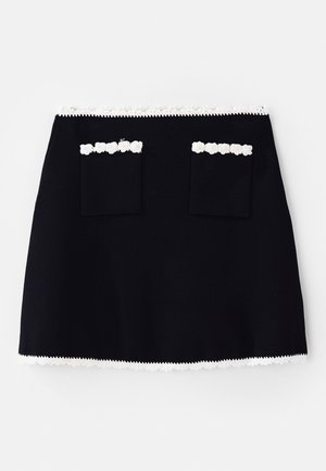 Sort nederdel med en hvid blondekant nederst, forstærket med to forlommer udsmykket med blonde detaljer. Glat tekstur, A-formet silhuet.