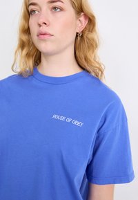Modré bavlněné tričko s krátkým rukávem, které má na levé straně hrudi bílé logo "HOUSE OF OBEY".