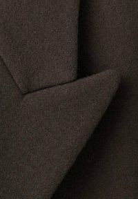 Tela de lana marrón oscuro y texturizada con una superficie suave similar al fieltro. Presenta esquinas superpuestas que crean un diseño angular y definido.