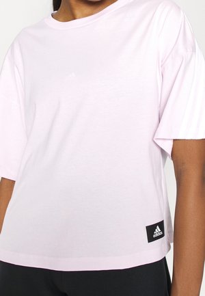 Kvinde iført en lyserød Adidas t-shirt med korte ærmer og et sort Adidas-logo tag nær den nederste kant.