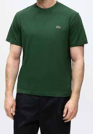 Mann trägt ein schlichtes, dunkelgrünes kurzärmeliges T-Shirt mit kleinem Krokodil-Logo auf der linken Brust und schwarze Hose.