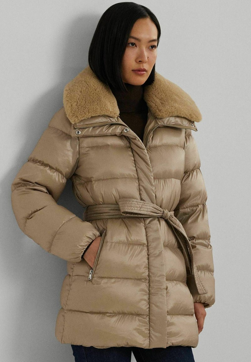 Beige gewatteerde jas met een nepbont kraag, een ceintuur in de taille en ritssluitingen aan de zijkanten. Kenmerkt zich door een gestikt oppervlak en een glanzende afwerking.