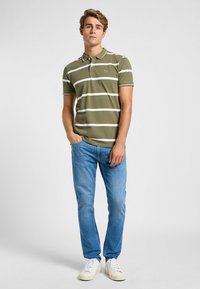 Polo verde oliva con rayas blancas, de manga corta y con cuello. Combinado con vaqueros azul claro y zapatillas blancas. Diseño simple y casual.