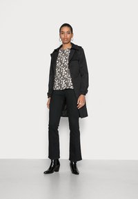 Casaco preto de trench coat sobre uma blusa floral, combinado com calças pretas largas e botas de salto pretas. Tecido suave e silhueta ajustada.