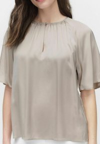 Beige blus tillverkad av ett mjukt tyg, korta ärmar, rund halsringning med rynkdetalj och en liten nyckelhålsöppning fram.