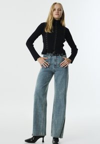 Un pull à col roulé noir côtelé avec des boutons à clous, associé à un jean large bleu clair avec des fentes sur les côtés et une ceinture noire. Bottes à talons noires.