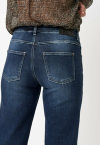 Des jeans en denim bleu foncé présentent un design classique à cinq poches, des coutures visibles, et une étiquette en cuir au niveau de la taille avec le détail de la marque au dos.