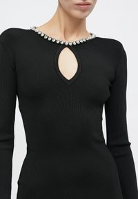 Γυναίκα που φοράει εφαρμοστή μαύρη μακρυμάνικη μπλούζα με κοψίματα σε σχήμα σταγόνας και κολιέ με μεγάλα διάφανα πολύτιμα πετράδια γύρω από το λαιμό.