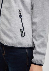 Šedá mikina na zip s námořnickými akcenty, vybavená boční zipovou kapsou a lemem z pružného materiálu. Hladký textilní povrch, přiléhavý střih.