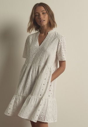 Mujer con vestido blanco de encaje de manga corta con escote en V y falda con volantes, de pie delante de un fondo liso y claro.
