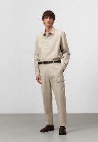 Uomo che indossa una tuta da lavoro beige con tasche laterali, cintura nera e scarpe marrone scuro, in piedi davanti a un muro bianco semplice.