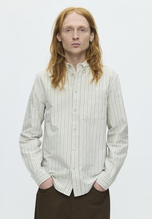 Jeune personne aux longs cheveux roux portant une chemise blanche à rayures grises avec des boutons et un pantalon foncé, debout les mains dans les poches.