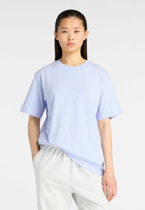 Giovane donna con lunghi capelli scuri che indossa una t-shirt azzurro chiaro a maniche corte e pantaloni grigio chiaro, con una mano in tasca.