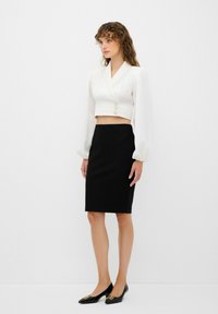 Blusa blanca corta con mangas abullonadas y tres botones dorados, combinada con una falda lápiz negra y zapatos negros de punta con detalles dorados.