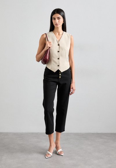 Gilet beige senza maniche con scollo a V e chiusura a bottoni, abbinato a pantaloni neri alla caviglia con bottoni dorati e sandali con tacco bianchi.
