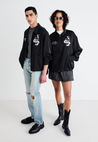 Črne zadrge jake s vezenimi motivi, oblečene čez sproščene outfite. Ena je kombinirana s svetlimi poderjenimi kavbojkami; druga pa z črtasto krilom in črnimi čevlji.