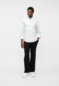 Άνδρας όρθιος με τα χέρια στις τσέπες, φοράει ανοιχτό πράσινο ριγέ πουκάμισο με κουμπιά, μαύρο παντελόνι και άσπρα αθλητικά παπούτσια σε απλό λευκό φόντο.