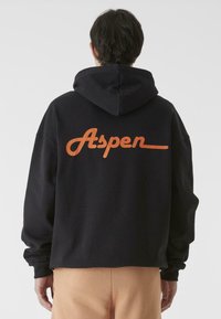 Fekete kapucnis pulóver puha anyagból, amelyen egy hangsúlyos narancssárga "Aspen" logó található a hátsó részen. Nagy méretű fazon, kenguruzsebbel és nagy kapucnival.