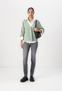 Svetlozelený rebrovaný cardigan s tromi gombíkmi, nosený cez bielu košeľu s golierom. Šedé slim-fit džínsy a čierne platformové mokasíny dopĺňajú vzhľad.