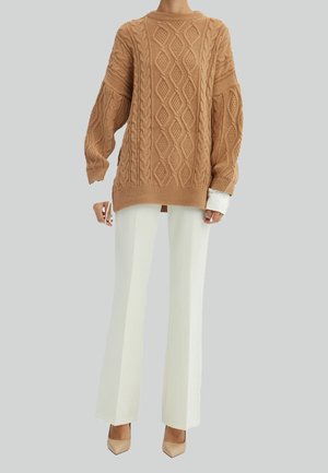 Kvinde iført en kamelfarvet, kabelstrikket oversized sweater, hvide bukser med vide ben og beige spidse hæle på en ensfarvet grå baggrund.