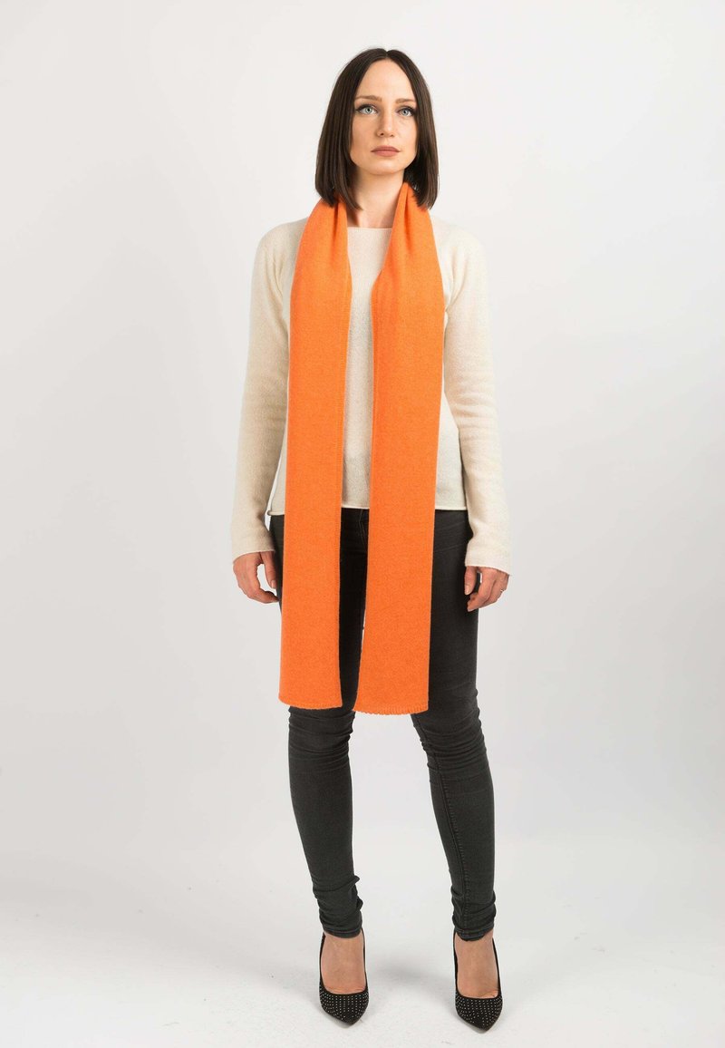 Orange uldescarf med en glat tekstur, draperet omkring halsen. Båret over en cremefarvet sweater og sorte skinny jeans, parret med sorte hæle.