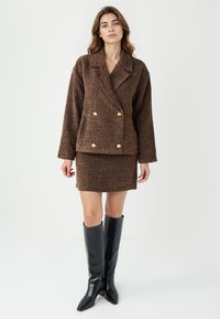 Bruine gestructureerde double-breasted jas met gouden knopen, gecombineerd met een bijpassende rok en kniehoge zwarte leren laarzen.