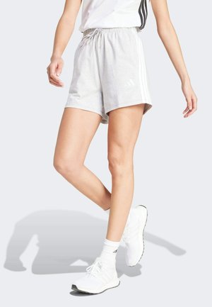 Personne portant un short de sport gris clair avec des rayures blanches et des baskets blanches, debout sur une jambe sur un fond neutre.