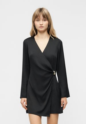 Ung kvinde med glat blondt hår iført en sort slå-om-kjole med lange ærmer og et gyldent spænde i højre side.