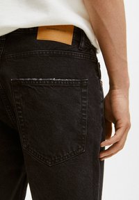 Jeans de mezclilla negra con un bolsillo trasero desgastado, que presenta una etiqueta de cuero marrón con la inscripción "PULL&BEARDENIM". Tejido texturizado con detalles de costura sutiles.