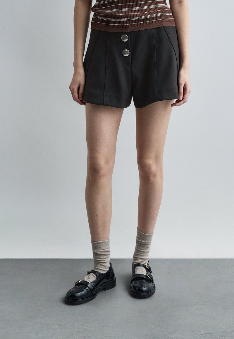 Γυναίκα που φοράει μαύρα σορτς με κουμπιά, μπεζ κάλτσες και μαύρα παπούτσια με αγκράφα, στέκεται σε γκρι πάτωμα μπροστά σε απλό φωτεινό τοίχο.