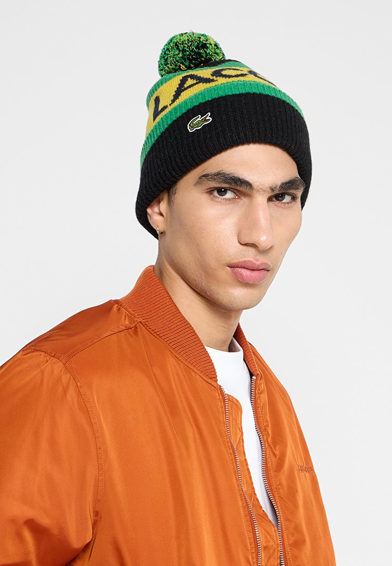 Gebreide muts in zwart, groen en geel met een pompon bovenop. Kenmerkt zich door een gedurfde "LAC"-ontwerp en een klein Lacoste-logo aan de zijkant.