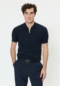 Giovane con capelli ricci che indossa una maglietta blu navy con zip a maniche corte e pantaloni a righe sottili, in piedi con una mano in tasca.