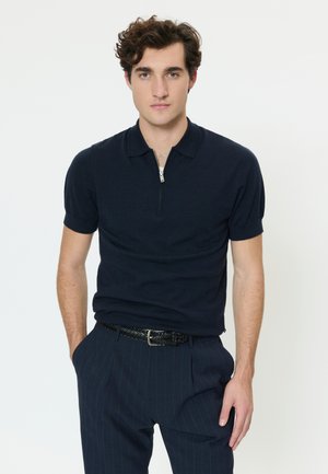 Jaunas vyras su garbanotais plaukais, vilkintis tamsiai mėlyną trumpomis rankovėmis su užtrauktuku marškinėlį ir kostiumines kelnes su dryželiais, stovintis viena ranka kišenėje.